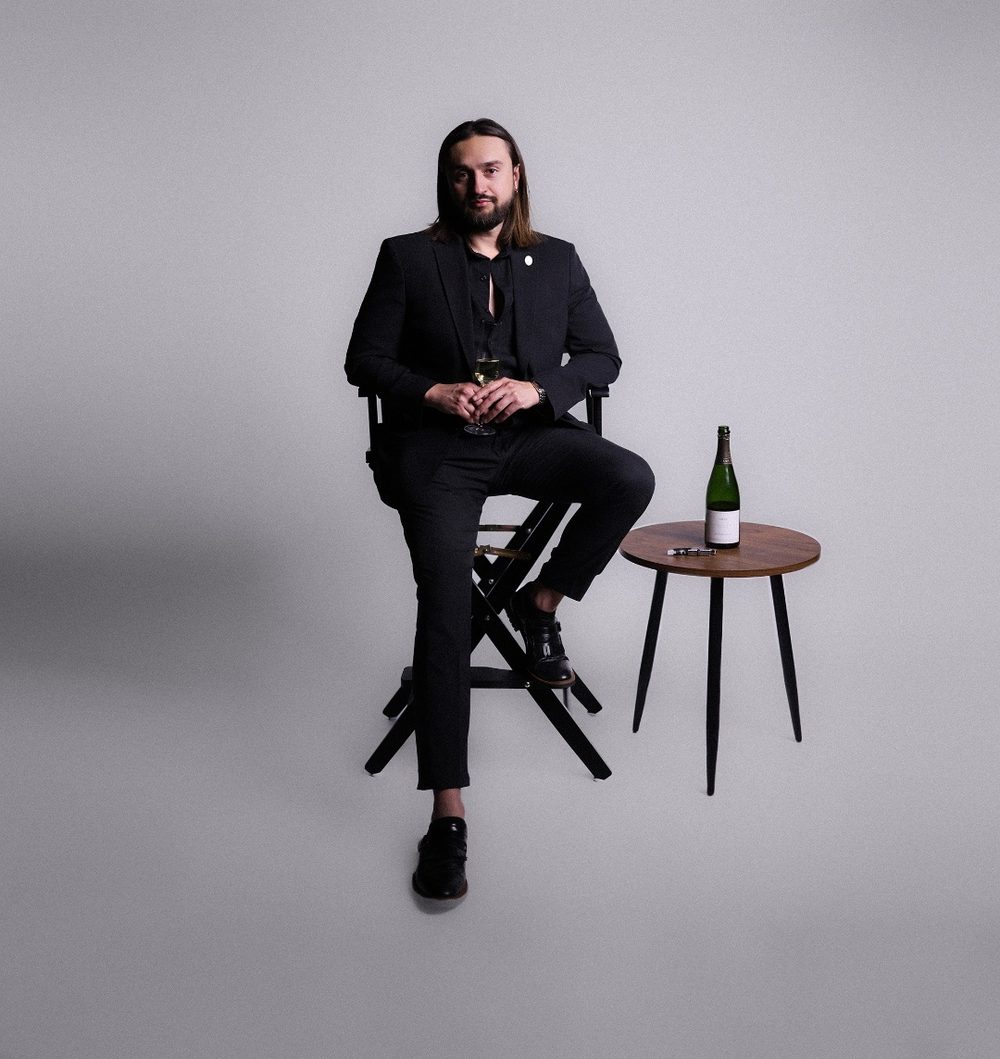 Nico Antuna Cooper seated on a director's chair beside a small table with a bottle of Champagne